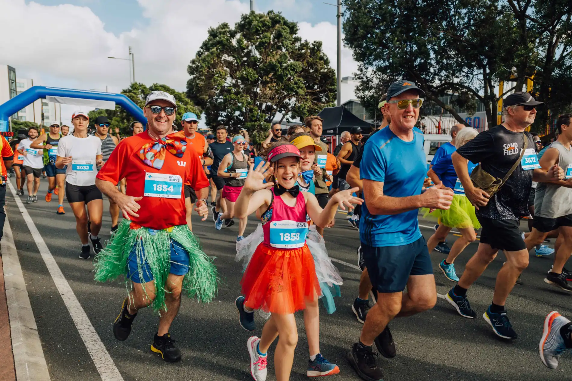 A group of people, including children and adults, are running in a race. They are wearing colorful outfits and tutus. The background shows a blue inflatable arch marking part of the race, and there are trees and spectators lining the route.