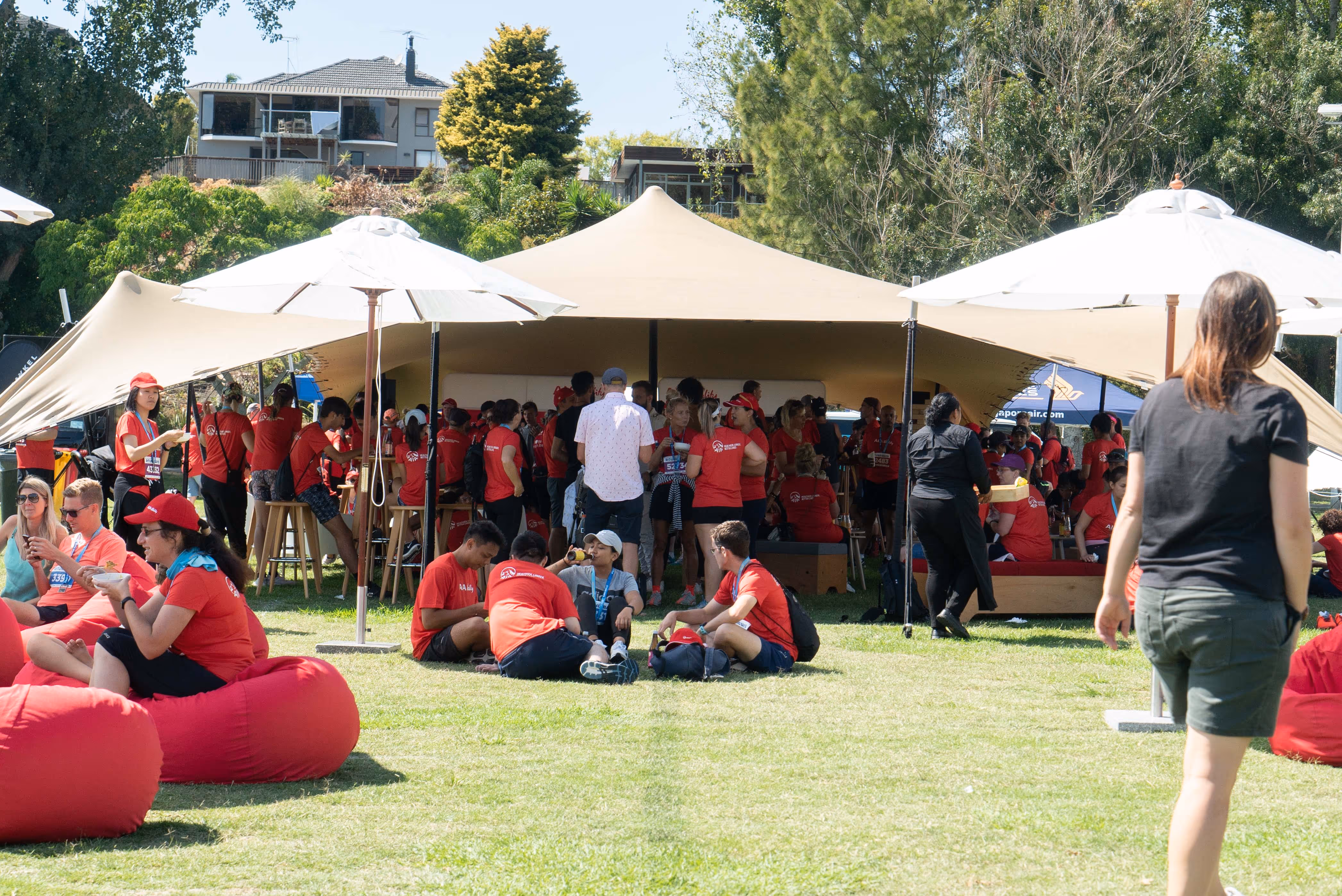 A group of people, many wearing red shirts, gather outdoors under large beige canopies. Some individuals sit on red bean bags on the grass, while others stand or walk around. Trees and a building are visible in the background on a sunny day.
