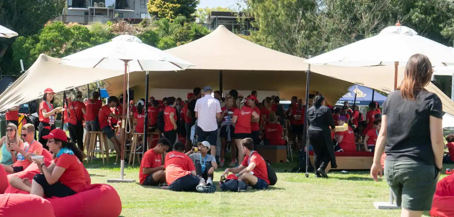 A group of people, many wearing red shirts, gather outdoors under large beige canopies. Some individuals sit on red bean bags on the grass, while others stand or walk around. Trees and a building are visible in the background on a sunny day.