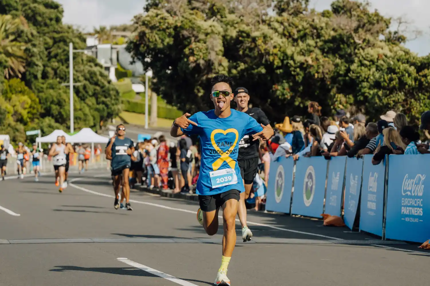 A runner in a blue shirt is crossing the finish line during a race, arms outstretched in celebration. Behind them, other participants and spectators are present, with trees and tents visible in the background.