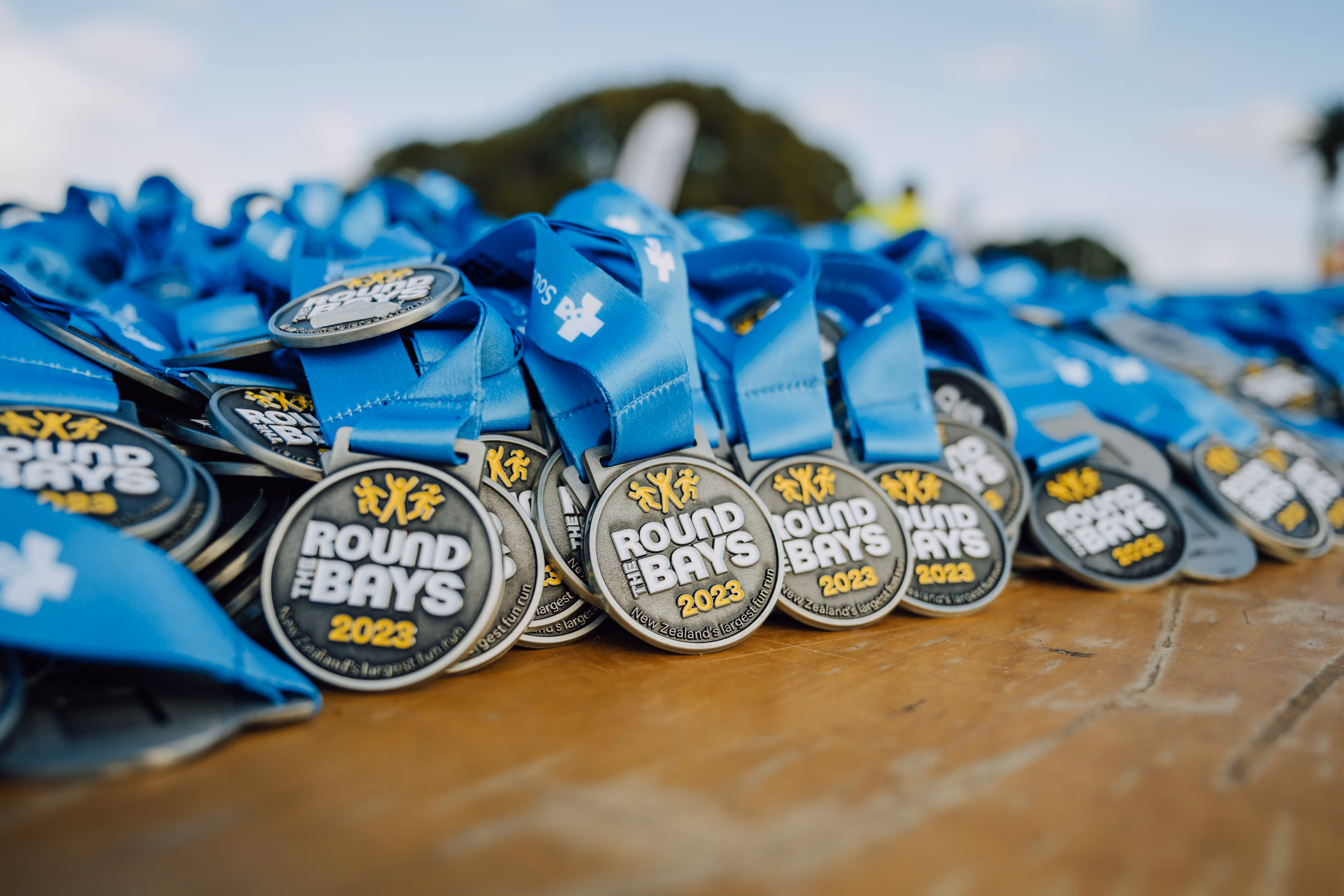 Close-up of several sports medals with blue ribbons laid out on a wooden table. Each medal reads "Round the Bays" in large text at the top and "2023" below. The background shows blurred greenery and people.