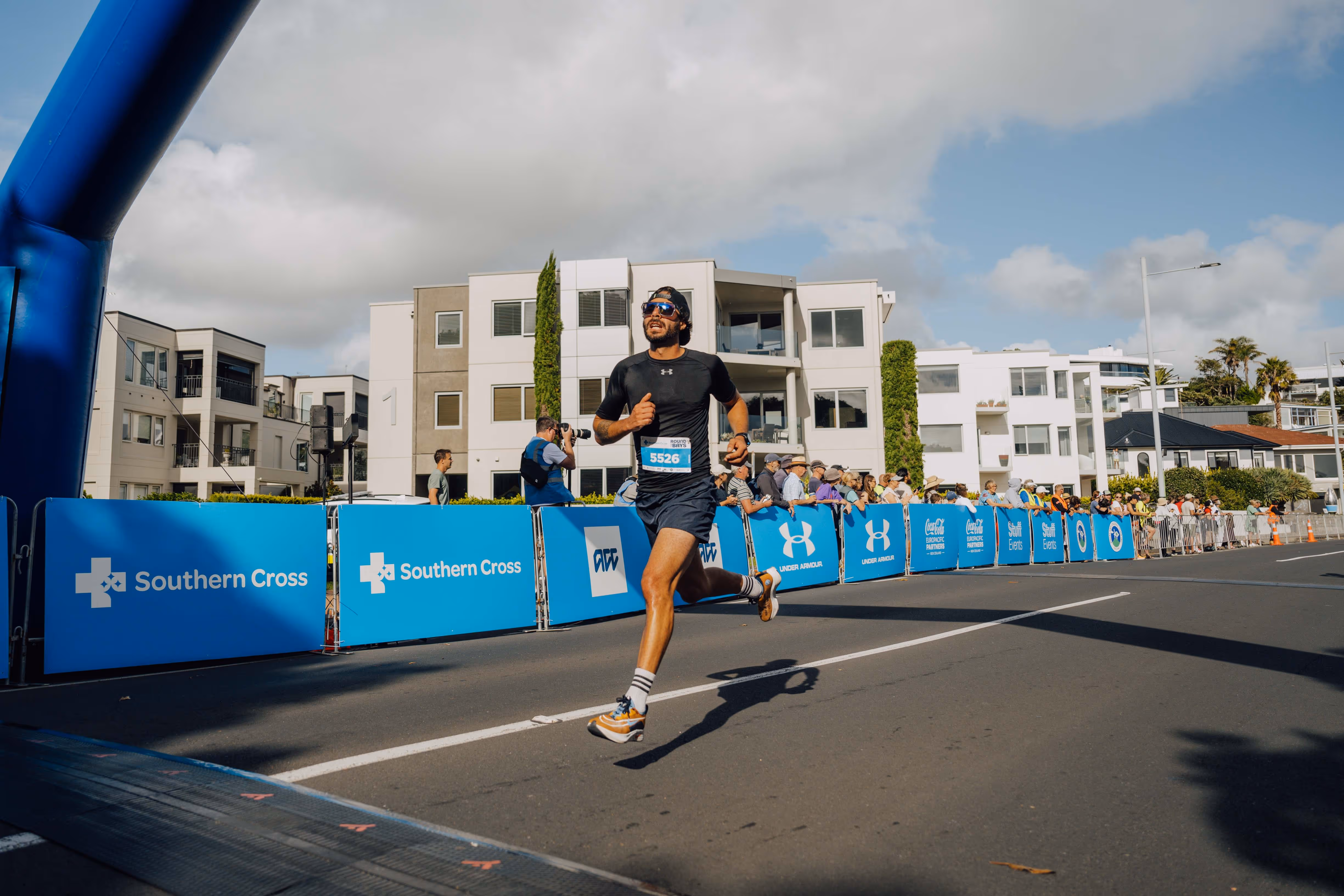 A person running towards the finish line of a race, with their foot crossing the line. The scene is outdoors, with spectators in the background and tall white buildings. Blue barriers with white logos line the race route. The sky is partly cloudy.