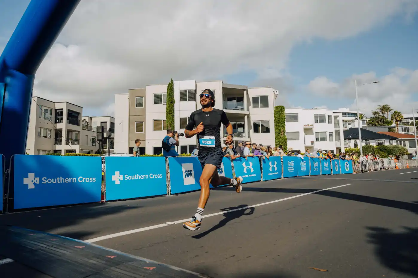 A person running towards the finish line of a race, with their foot crossing the line. The scene is outdoors, with spectators in the background and tall white buildings. Blue barriers with white logos line the race route. The sky is partly cloudy.