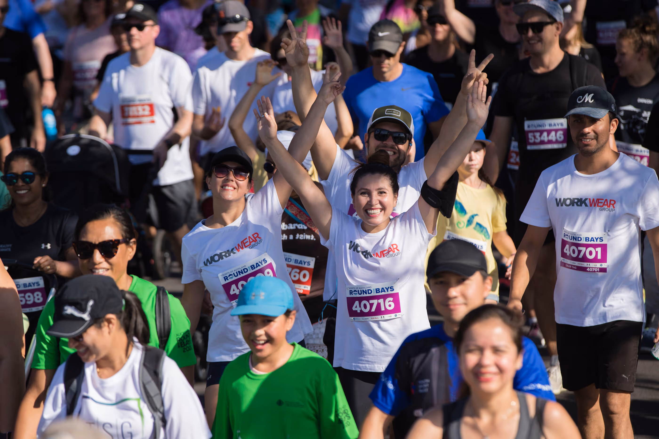 A large group of runners is gathered at the starting line of a race. The blue archway marking the "START" is prominently displayed. The scene is bustling with colorful attire and the road ahead is packed with participants. Trees and buildings are visible in the background.