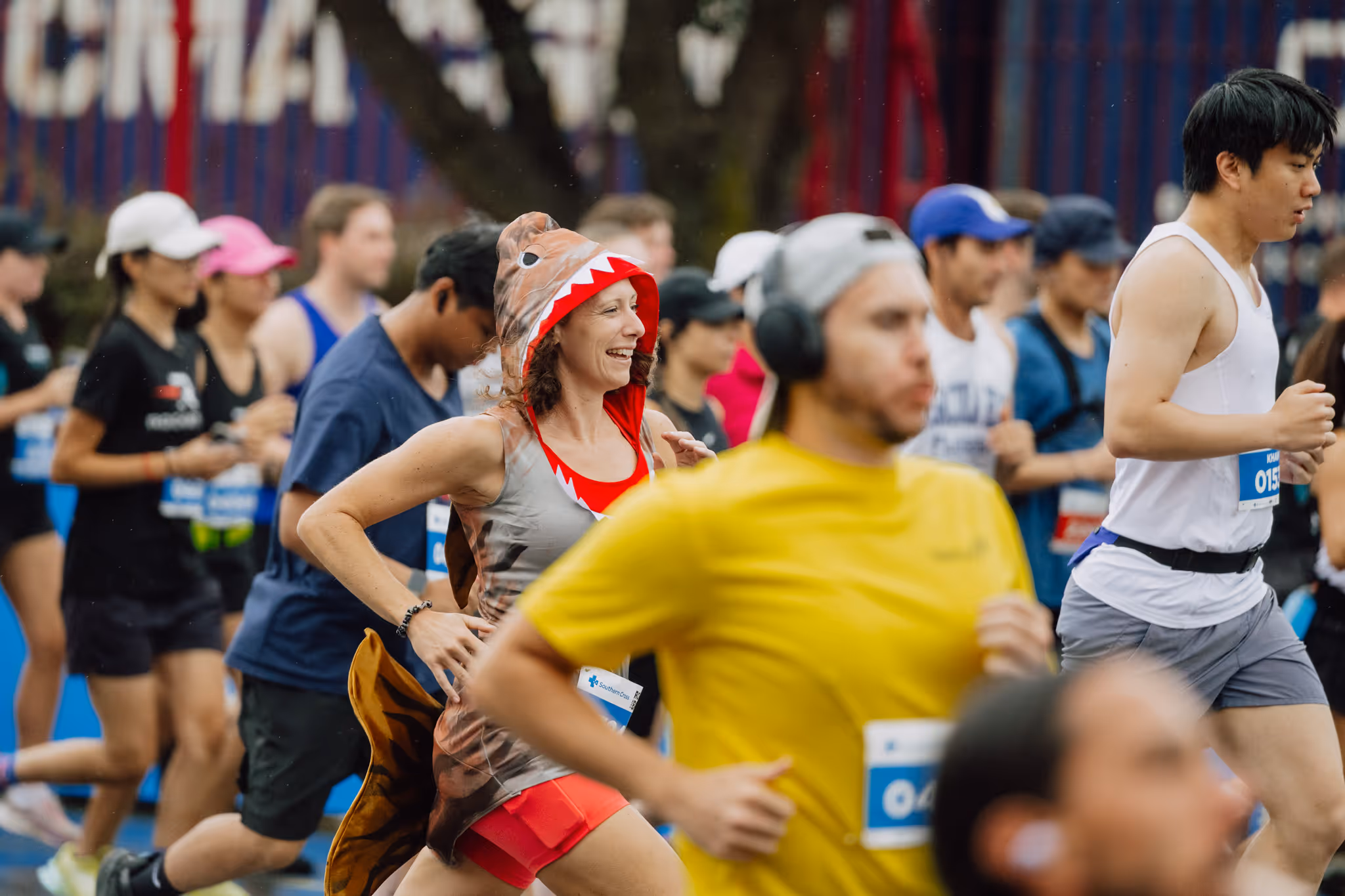 A group of runners of various ages participating in a race. The focus is on a woman in red and a man in yellow. Some wear headphones, hats, and wristbands while running. The background shows more runners and parts of trees and event signage.