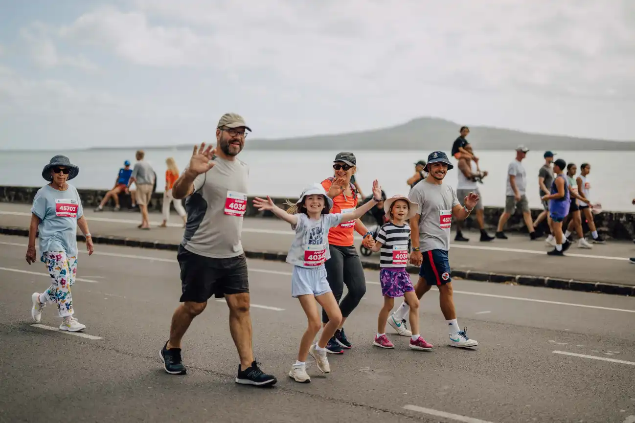 A group of people, including adults and children, walk together on a road by the seaside. The children hold hands and smile, while the adults wave. The background features a view of the ocean and a distant mountain under a cloudy sky. Other people walk nearby.