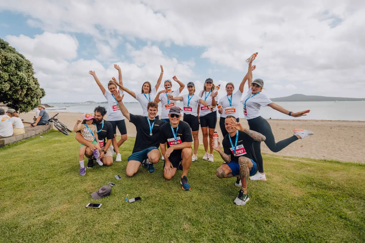 A group of people, some wearing medals, are joyfully posing with their arms raised, smiling, and displaying lively gestures on a grassy area by the beach. The background features a cloudy sky, distant water, and hills.
