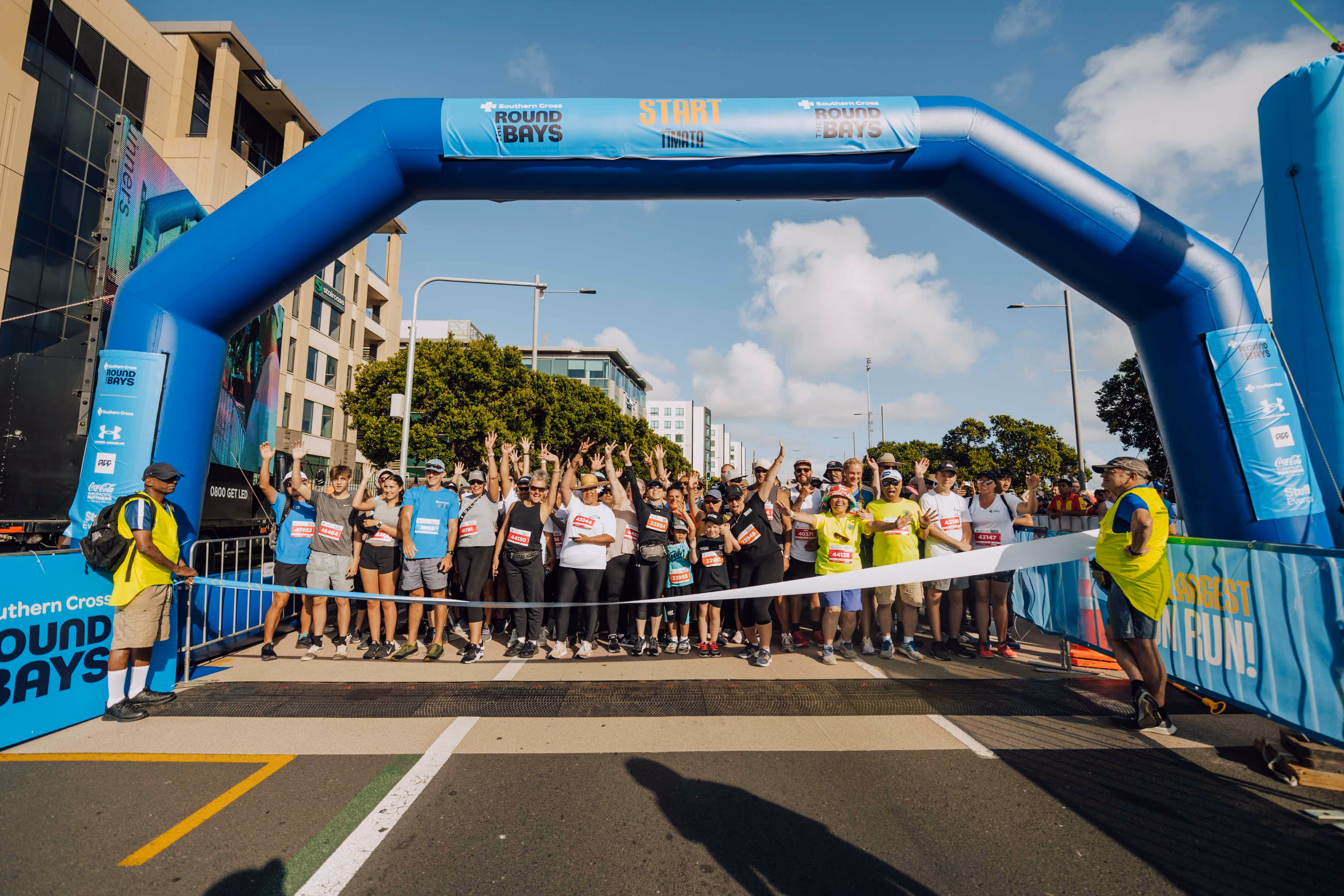 A large group of runners is gathered at the starting line of a race. The blue archway marking the "START" is prominently displayed. The scene is bustling with colorful attire and the road ahead is packed with participants. Trees and buildings are visible in the background.