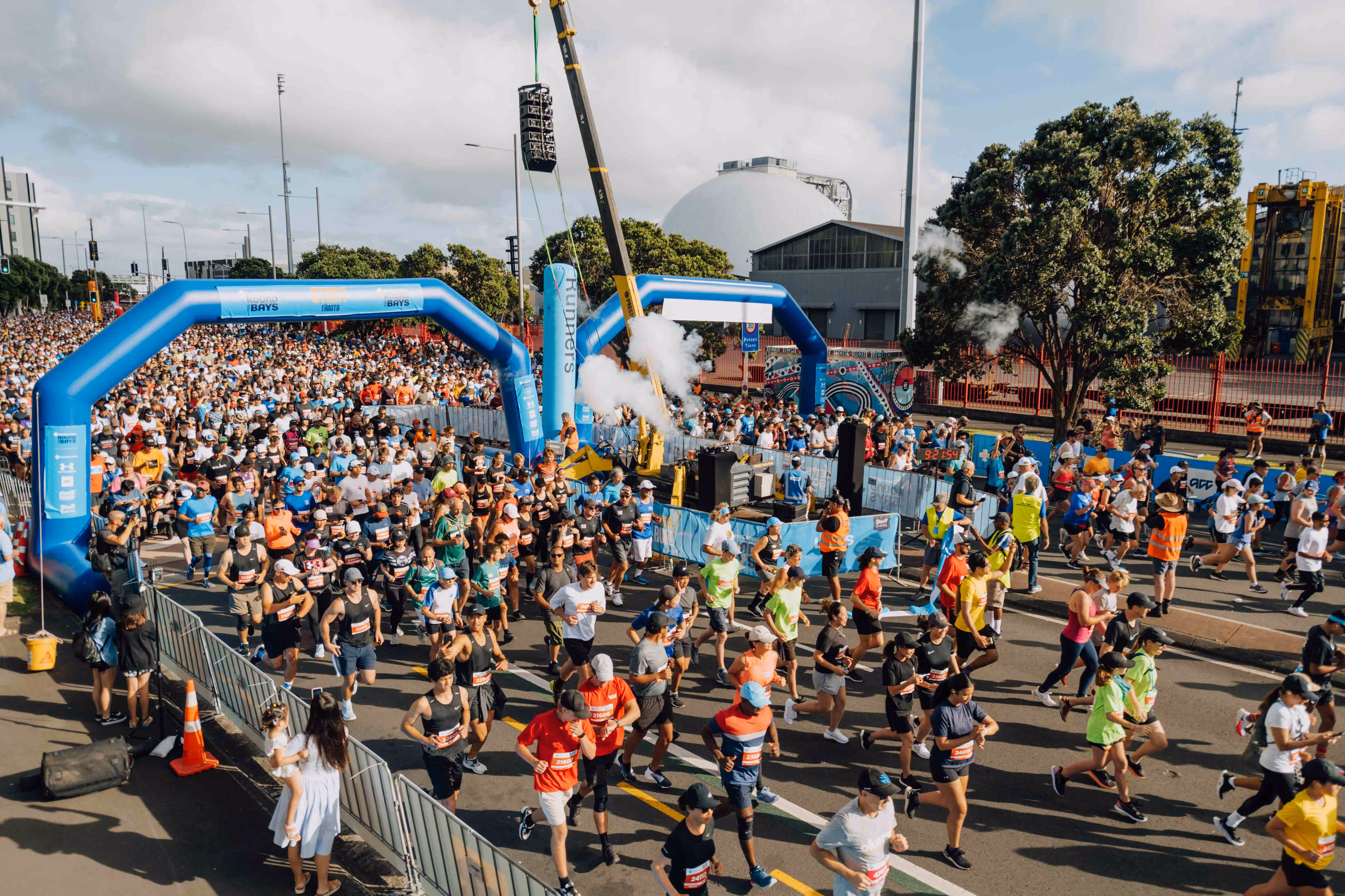 A large group of runners is gathered at the starting line of a marathon. The participants are dressed in various athletic gear and appear ready to begin the race. There are blue inflatable archways marking the start, and a crowd of spectators is visible in the background.