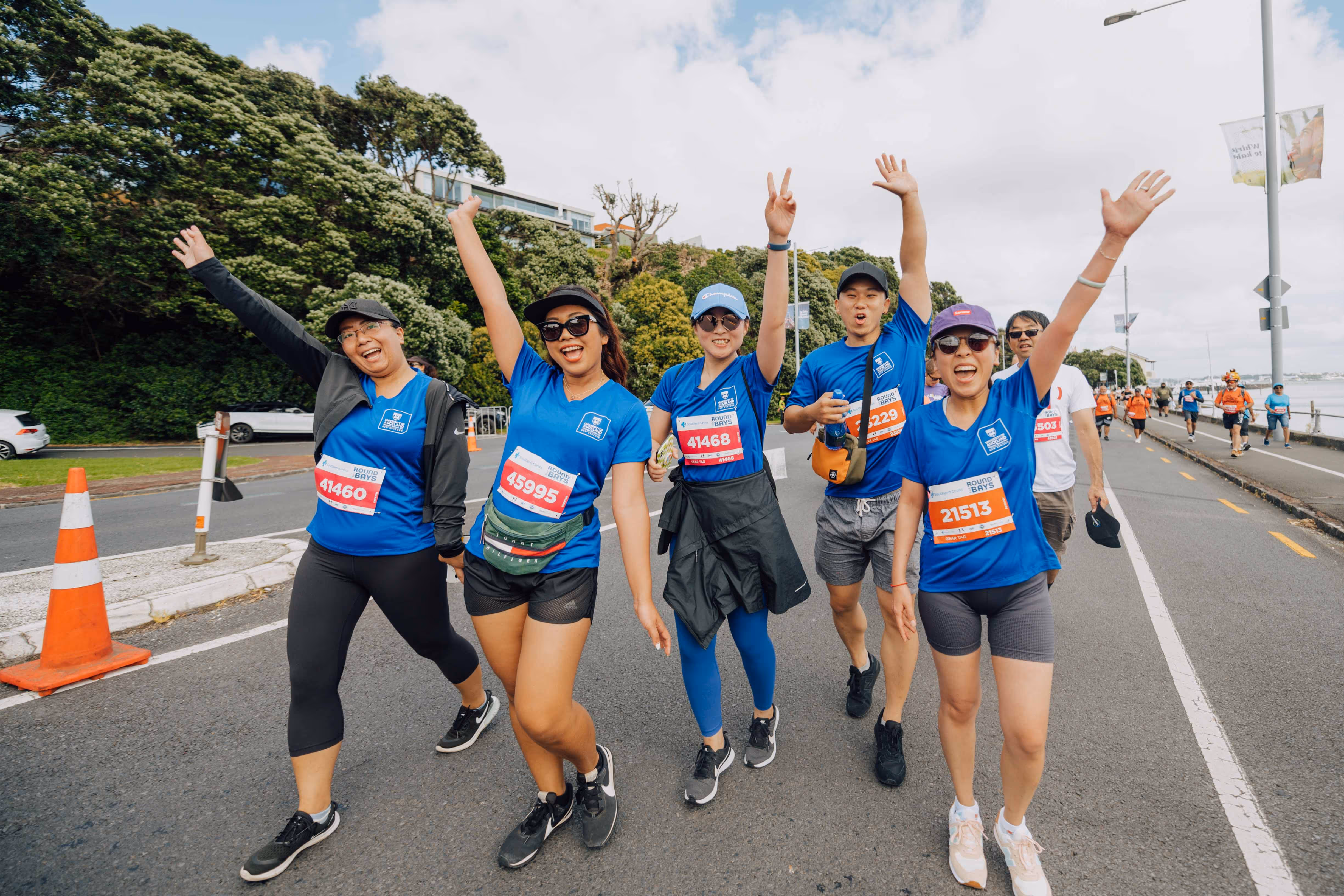 A group of six people wearing blue shirts and race bibs walk along a road with their arms raised in celebration. They appear to be participating in a race or charity walk. Traffic cones and greenery are visible in the background.