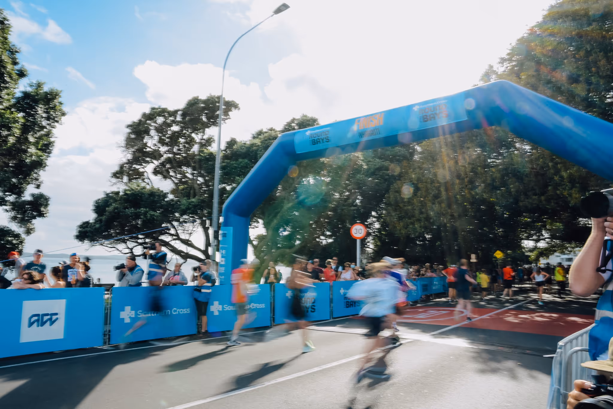 Runners cross the finish line at an outdoor race event. The finish is marked by a blue inflatable arch with trees and spectators in the background. Bright sunlight illuminates the scene, casting long shadows on the pavement.