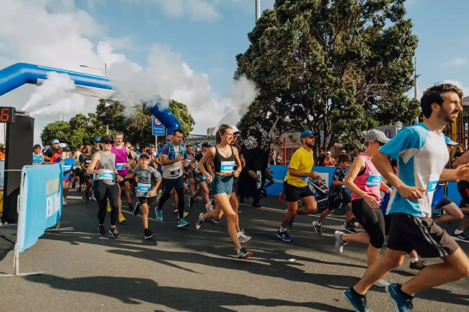 A group of runners participating in a road race, with a blue and white inflatable arch in the background. Some participants are wearing race bibs and athletic attire. Trees and spectators line the course on a sunny day.
