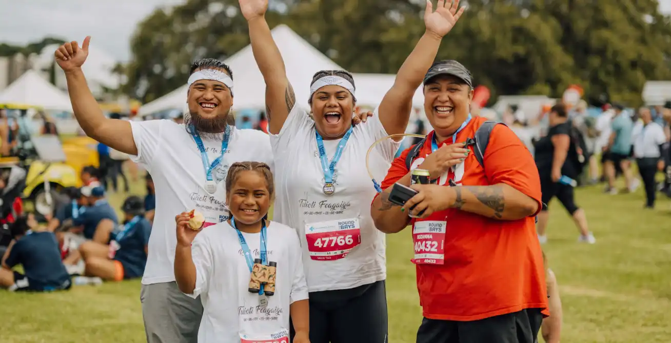Four individuals stand in a grassy area, celebrating after a race. Three people in white shirts and medals cheer with their arms raised, while one in a red shirt smiles and holds a camera. A child in front holds up a medal, and people and tents are visible in the background.