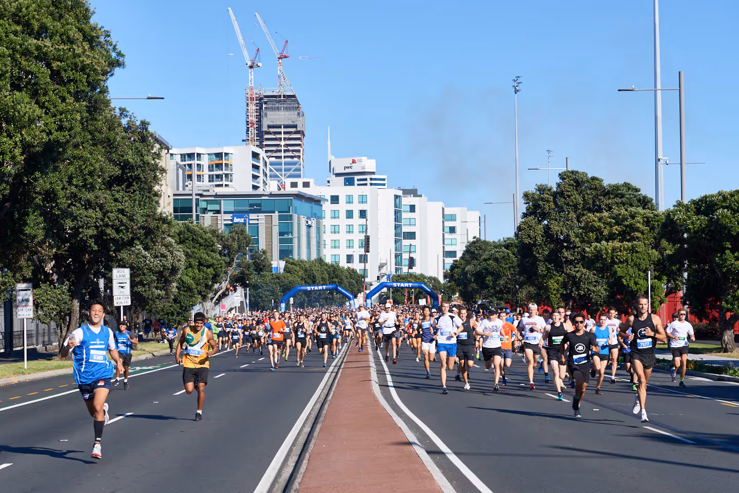A group of runners participating in a road race on a wide, divided city street. Tall buildings, cranes, and trees are visible in the background. Spectators and event signage line the course, and the weather appears clear and sunny.
