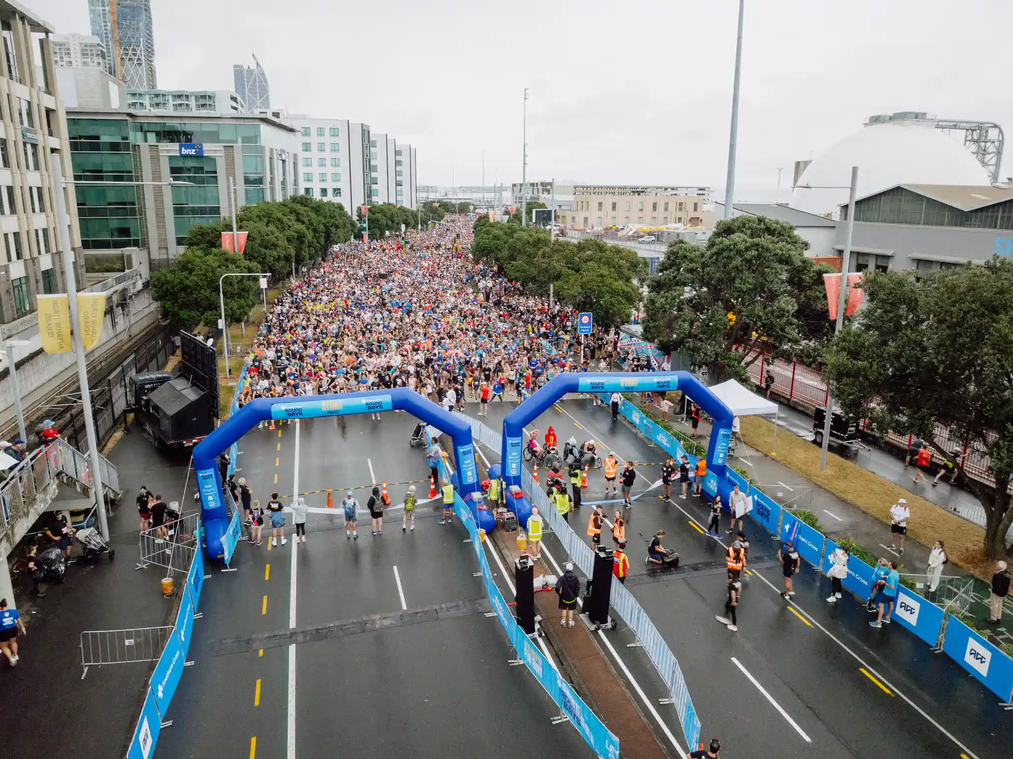 A large crowd of marathon runners is taking off from the starting line in a city. The race route is lined with blue and white barriers, and spectators watch from the sides. Buildings and trees are visible in the background under a cloudy sky.