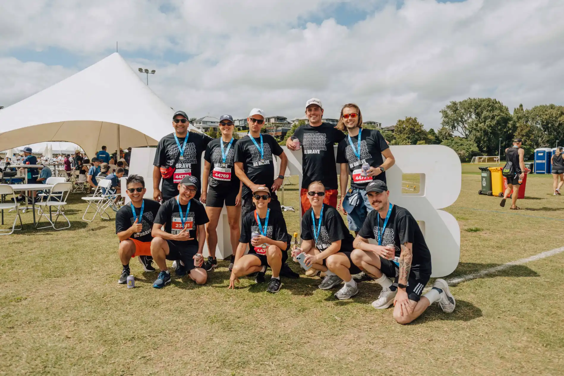 A group of ten people dressed in coordinated athletic wear poses for a photo outdoors near a large tent and a "B" sign. Some are standing, while others are crouching. The sky is partly cloudy, and the scene suggests participation in an event or competition.