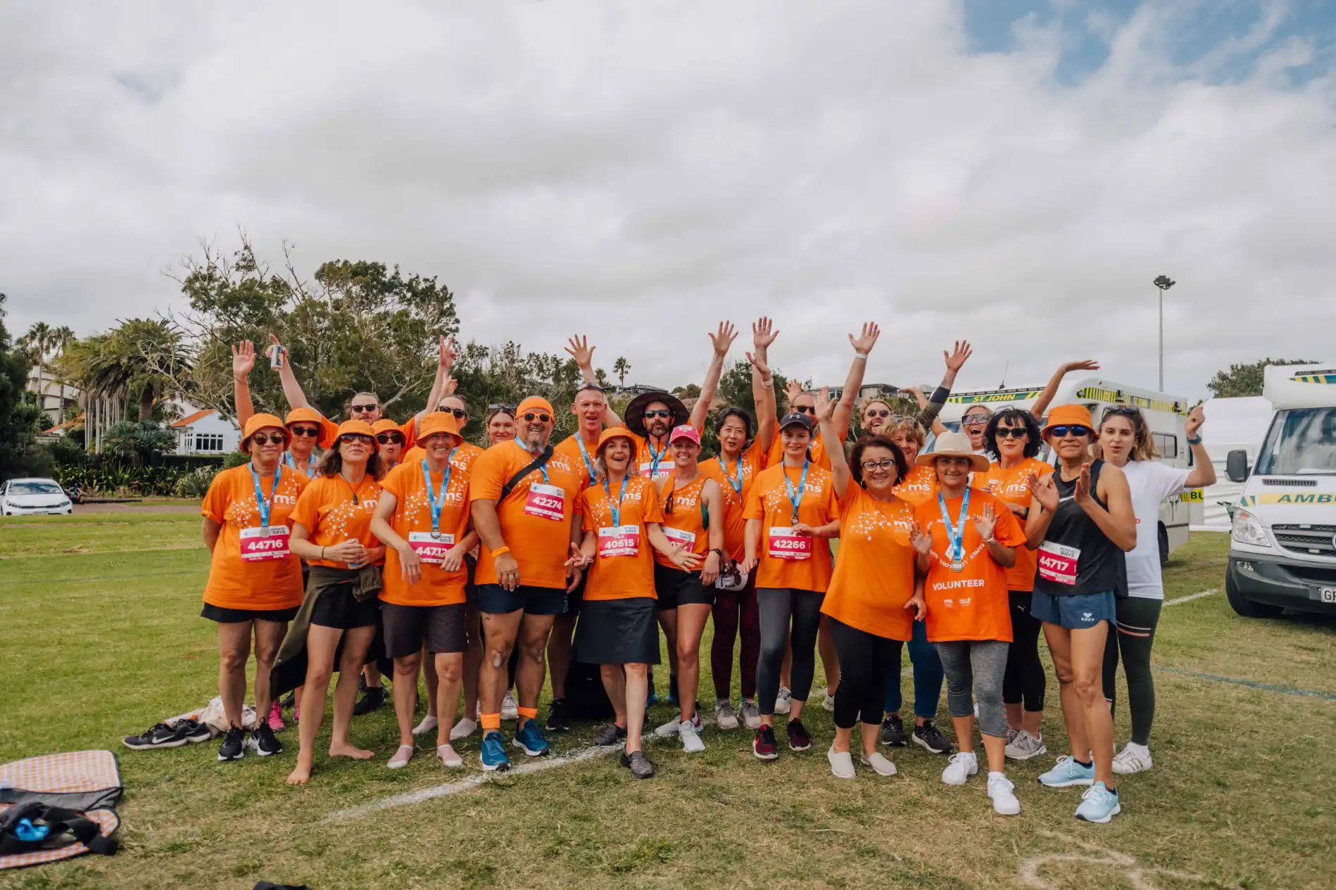 A group of people wearing orange shirts and holding drinks stand together outdoors on grass, several raising their arms in celebration. Many wear hats and sunglasses. Trees and vehicles are visible in the background, and the sky is partly cloudy.
