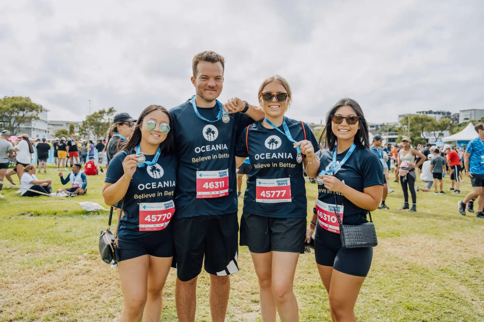 A group of four people wearing matching T-shirts and medals stands together outdoors at a community event or race. The group, which consists of three women and one man, smiles for the camera while people and tents are visible in the background.