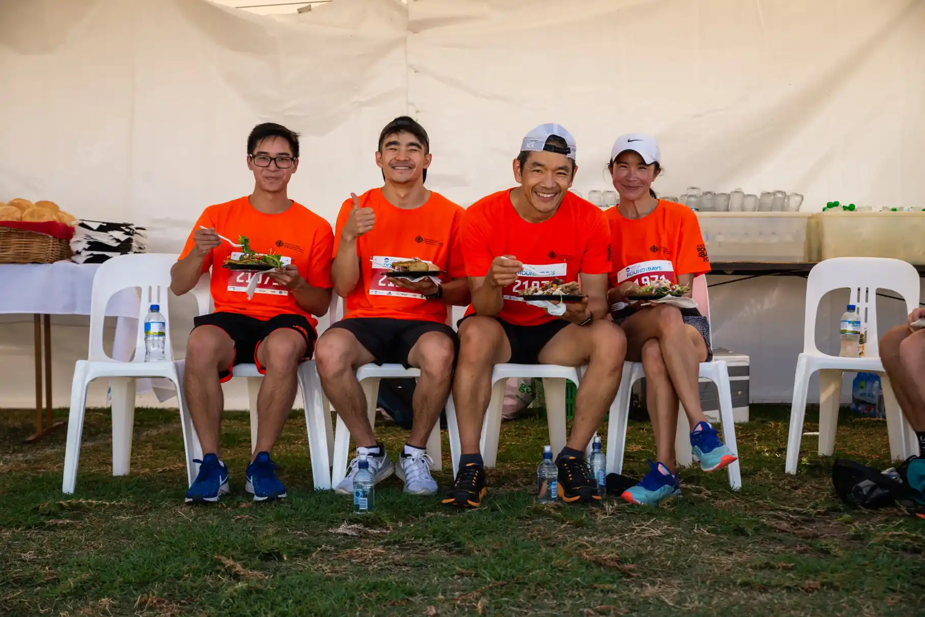 Four people in red tshirts and sports gear sit on white plastic chairs eating off plates and smiling at the camera