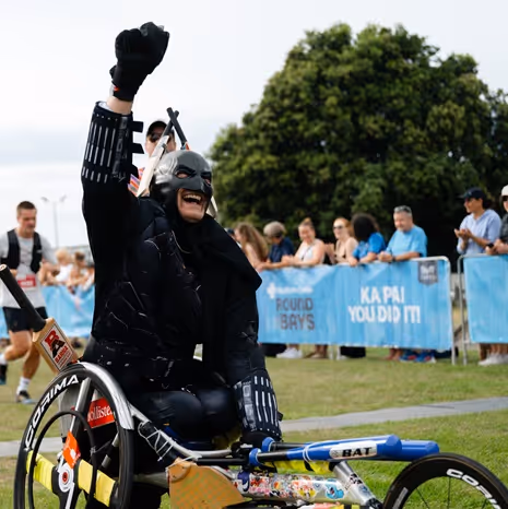 Participants, including wheelchair athletes, gather at the starting line of a race on a wet street. They are surrounded by supporters and are poised to start, with buildings and trees in the background.