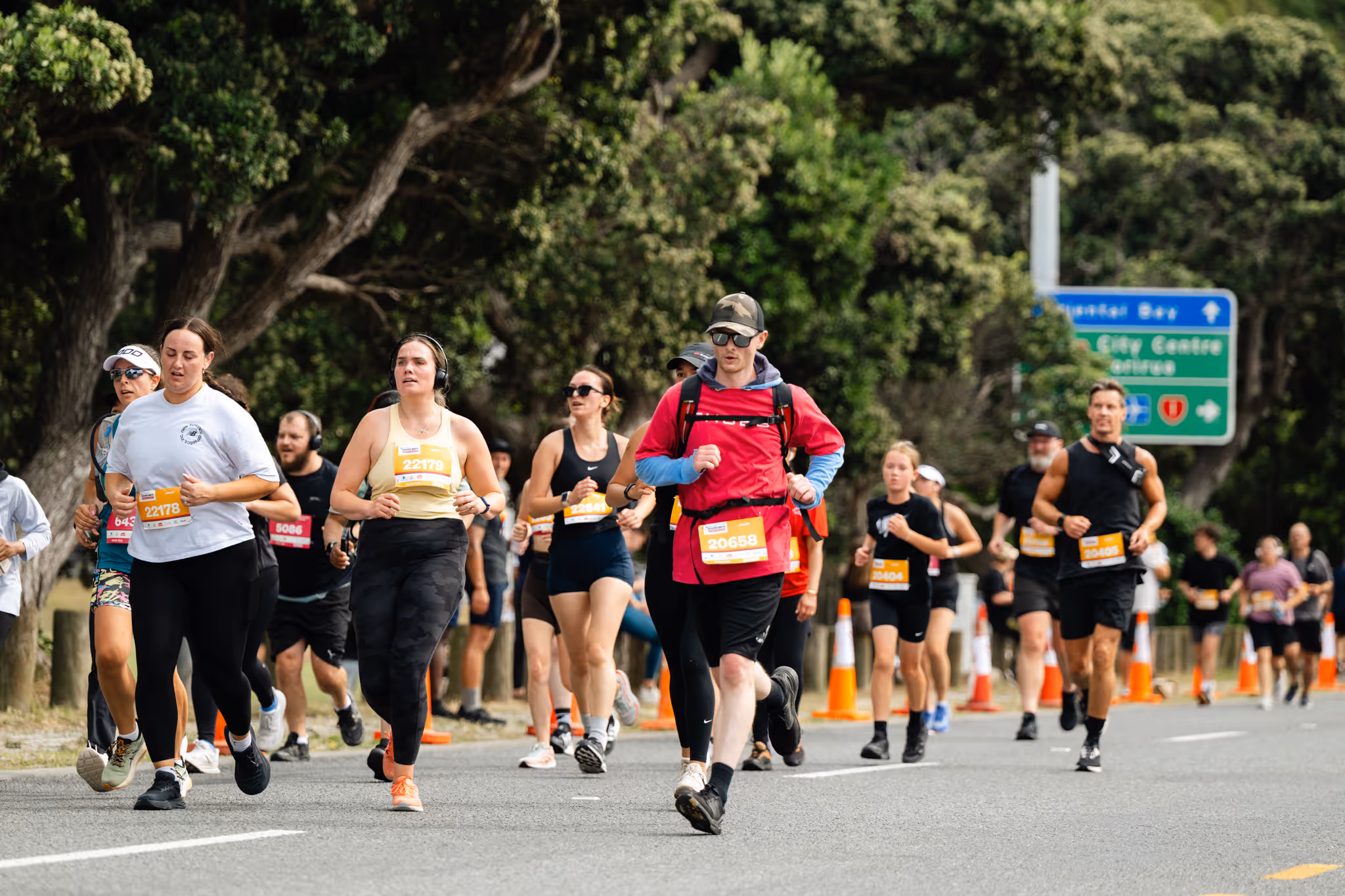 A large group of runners, including men and women, participates in a marathon along a seaside road. The sky is overcast, and there are hills and buildings visible in the distance across the water. The road has light posts, and some people watch from the sidelines.