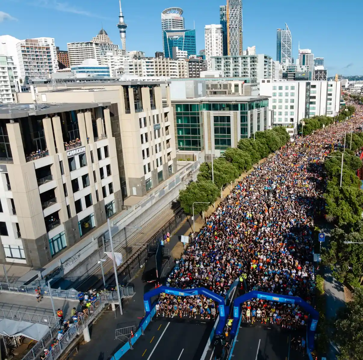 Two people in pink tutus run on a road during a race or fun run event. Both are smiling and have their arms raised in a celebratory manner. Other participants and spectators can be seen in the background. It's a sunny day with a few clouds in the sky.