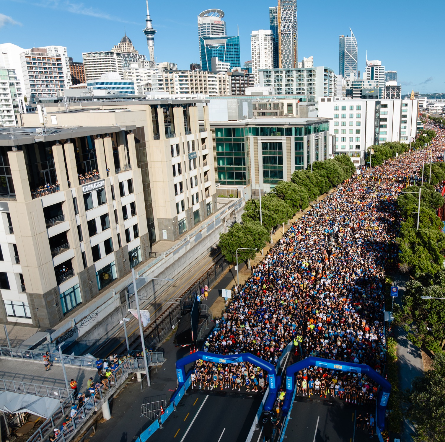 Two people in pink tutus run on a road during a race or fun run event. Both are smiling and have their arms raised in a celebratory manner. Other participants and spectators can be seen in the background. It's a sunny day with a few clouds in the sky.
