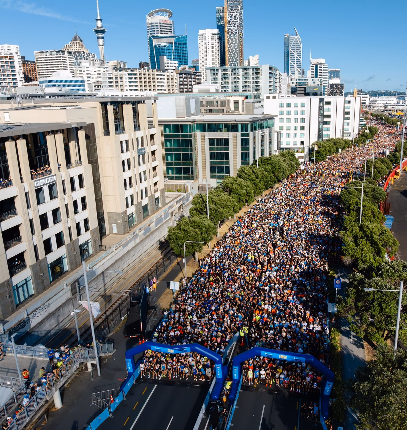 A large crowd of runners is gathered at the starting line of an outdoor marathon event. Blue inflatable arches mark the start, with streets lined by barriers and volunteers in orange vests. Surrounding the scene are tall city buildings and trees, under an overcast sky.