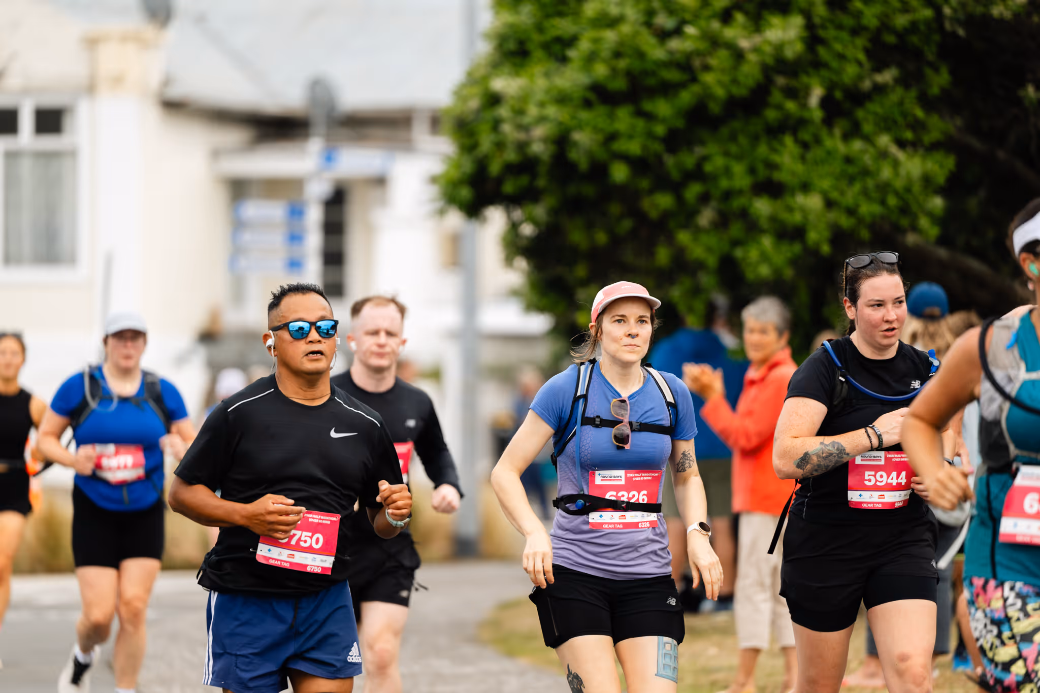 A runner in a blue shirt is crossing the finish line during a race, arms outstretched in celebration. Behind them, other participants and spectators are present, with trees and tents visible in the background.