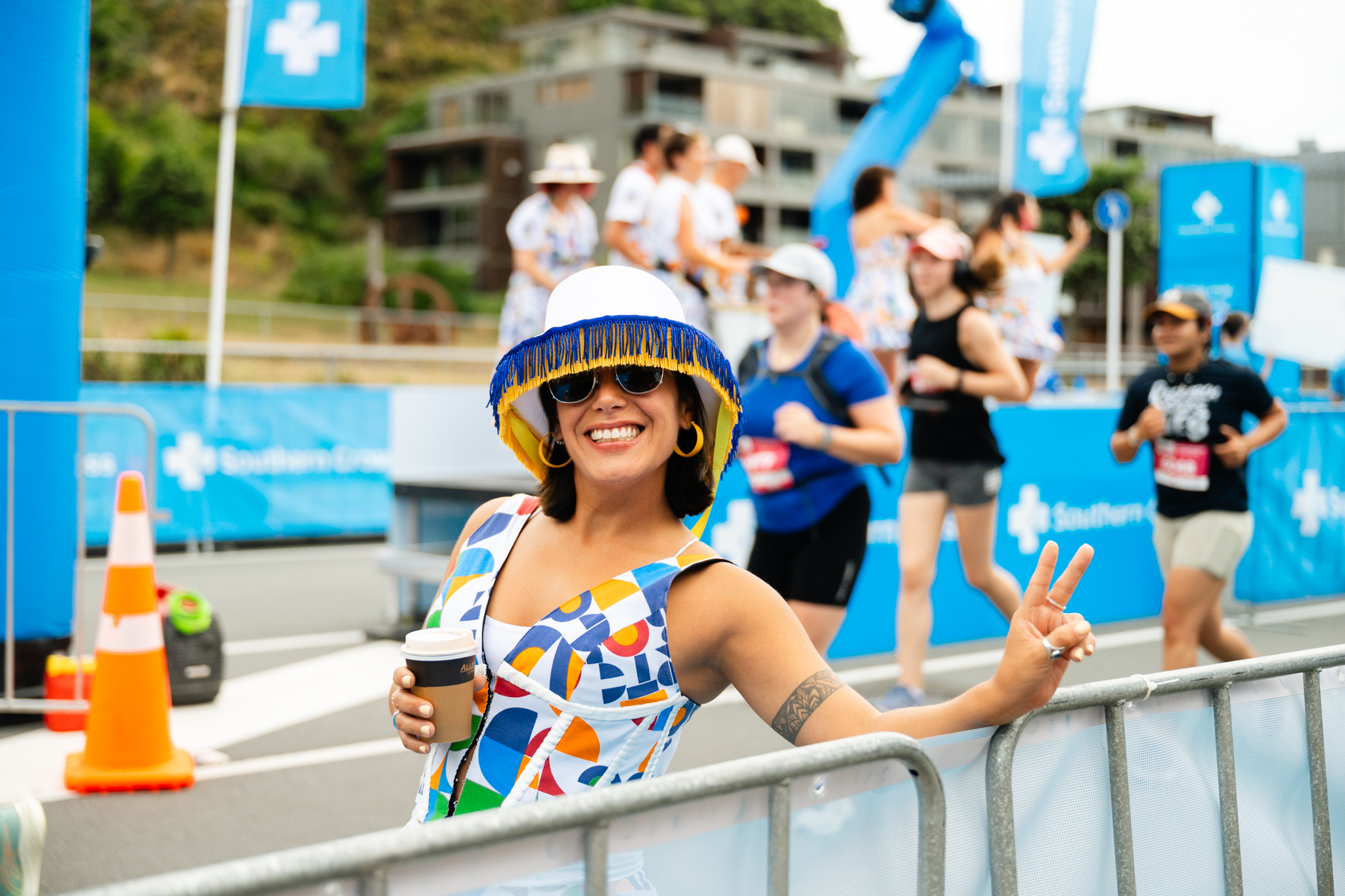 Three participants in yellow shirts celebrate with high-fives as they cross the finish line of a race. The finish line banner is blue and white. Spectators line both sides of the dirt track, cheering the runners. The sky is partly cloudy.