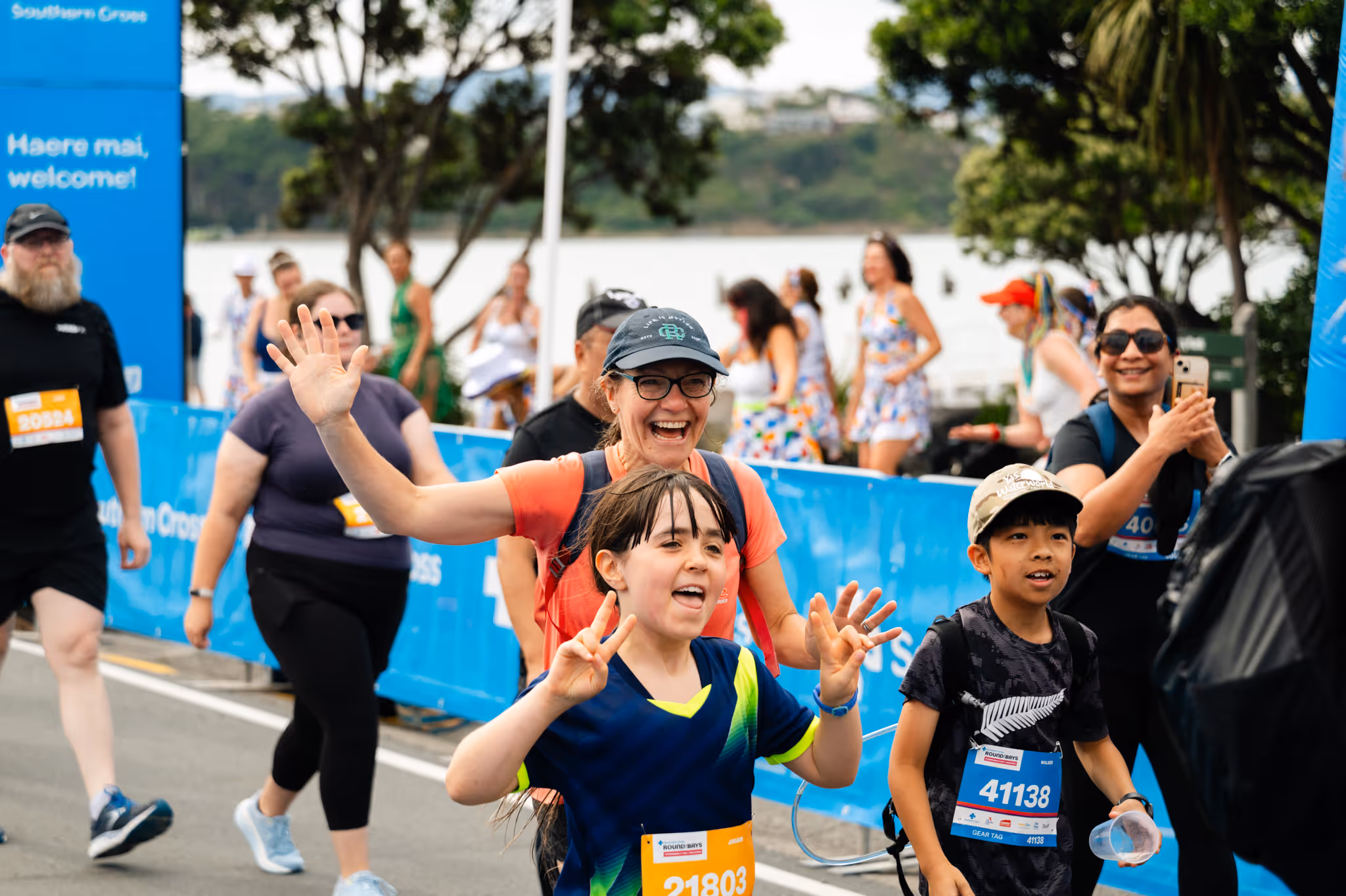 Two women in athletic gear cross the finish line of a race, holding hands and raising their arms in celebration. They are surrounded by cheering spectators and banners that say "Sport Wellington." The atmosphere is jubilant, and both women appear elated.