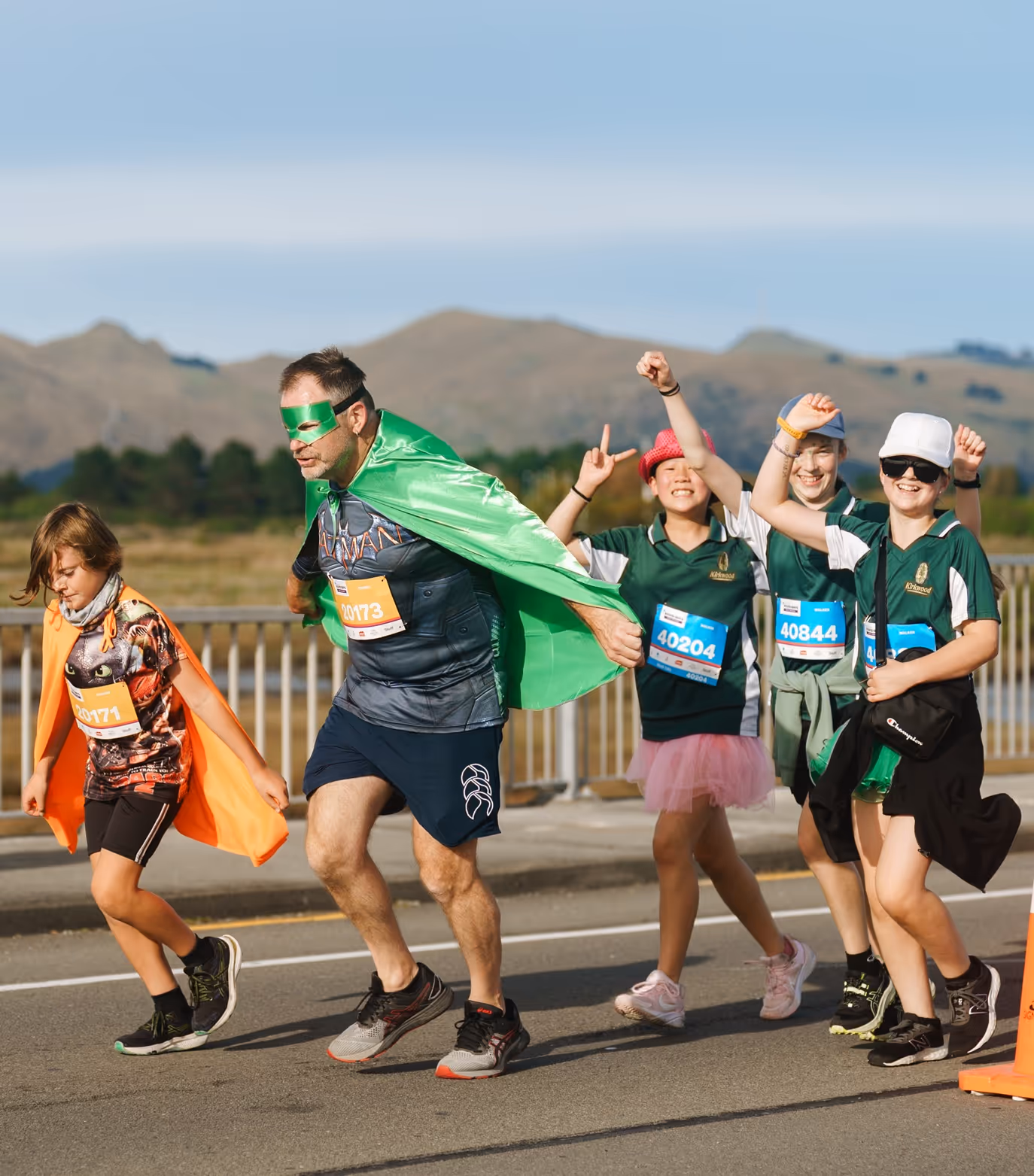 Two women in athletic gear cross the finish line of a race, holding hands and raising their arms in celebration. They are surrounded by cheering spectators and banners that say "Sport Wellington." The atmosphere is jubilant, and both women appear elated.