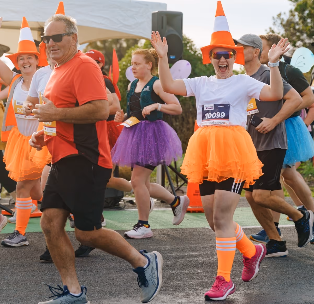 A group of runners of diverse ages and genders participate in an outdoor race on a sunny day, with two women in the foreground smiling, wearing athletic gear, and waving at the camera. The background shows other participants and trees.