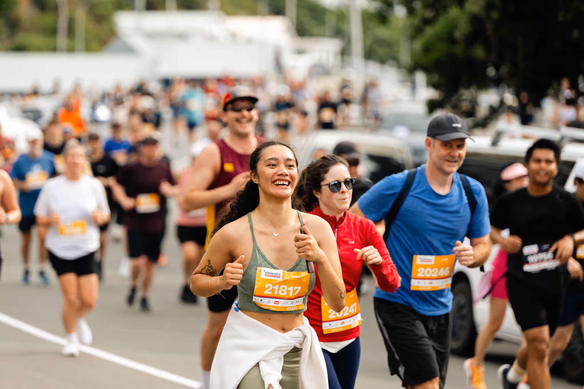 Three people holding hands and smiling while running in a race. The person on the left is wearing a tank top, the person in the middle is in a green shirt, and the person on the right is dressed in a blue shirt and shorts. Spectators and other runners are in the background.