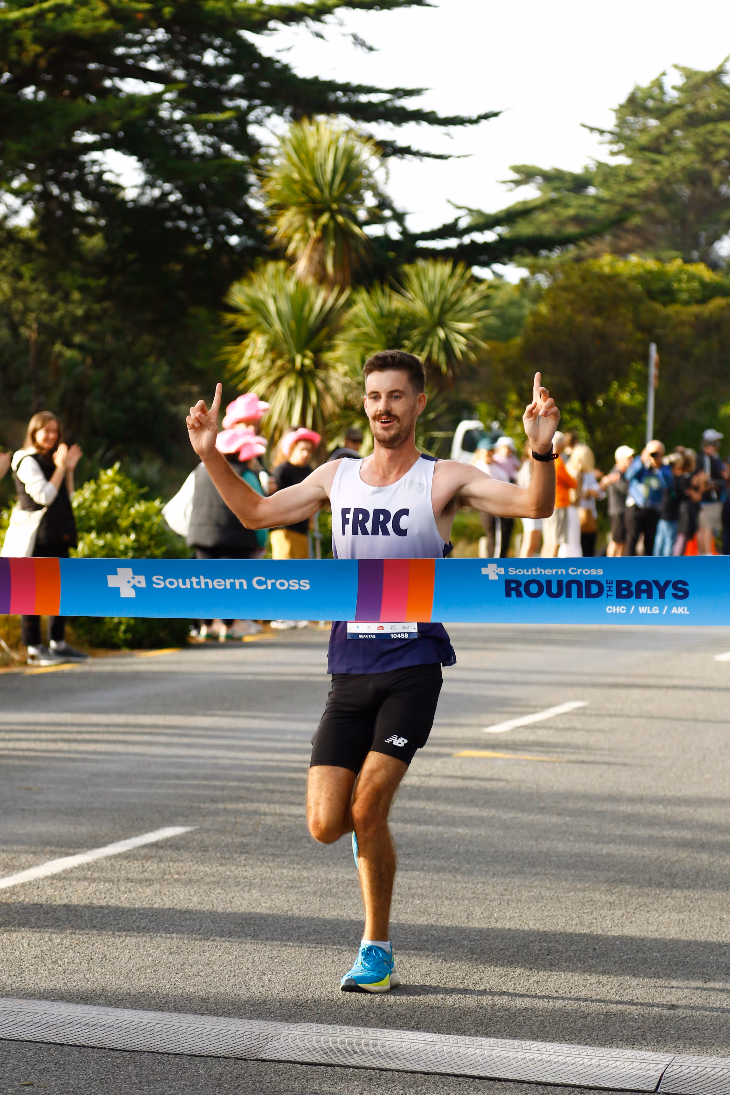 A woman wearing a black tank top and race bib smiles and makes a peace sign with both hands while running in a crowded race. Other participants are visible in the background on a paved road.