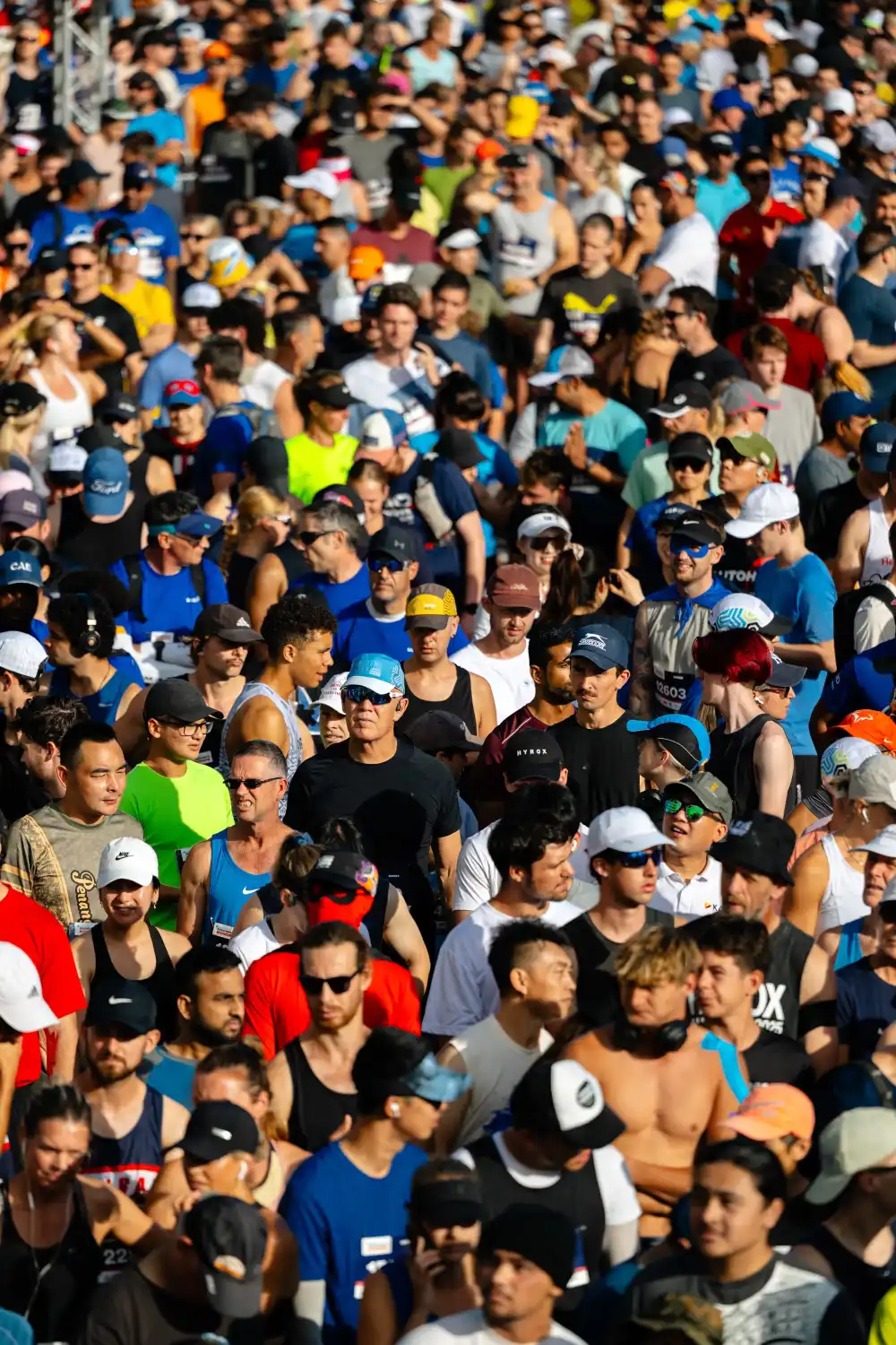 A large group of runners is gathered at the starting line of a race. The blue archway marking the "START" is prominently displayed. The scene is bustling with colorful attire and the road ahead is packed with participants. Trees and buildings are visible in the background.