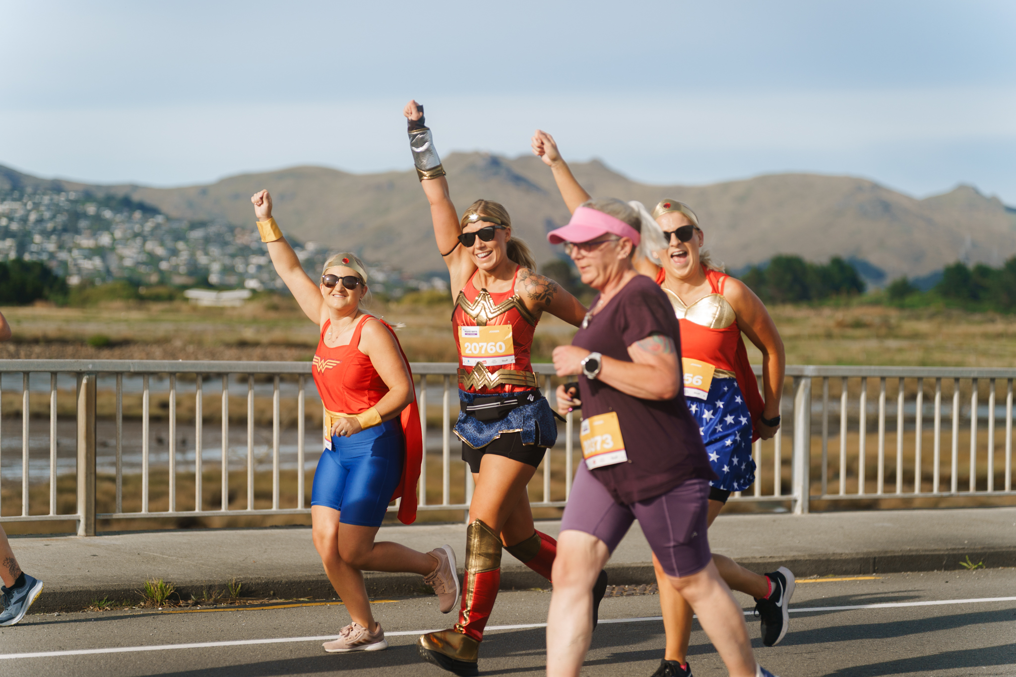 A smiling woman wearing a white shirt, black shorts, and a grey cap raises her arms in celebration during a road race. She has a race bib labeled "20176." Other participants, wearing various athletic clothing and bibs, are also running along the wet road. Trees are visible in the background.