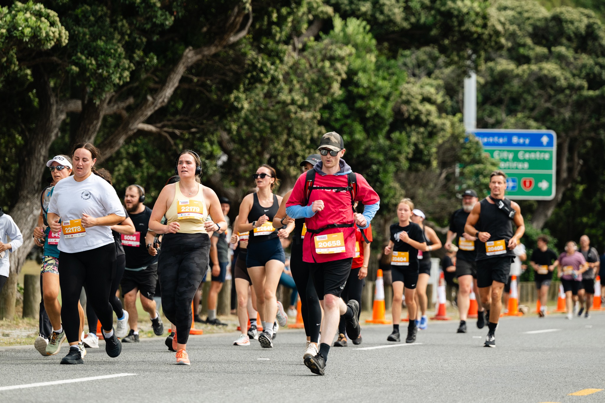 A group of runners in athletic gear competes in a marathon on a sunny day. They are running on a paved road flanked by green trees, with some road signs and traffic lights visible in the background. The runners are focused and appear to be mid-race.