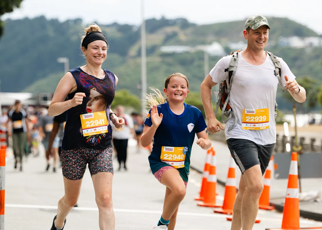 Three people wearing race bibs smile as they run together outdoors during a race, with orange cones lining the road and hills visible in the background.