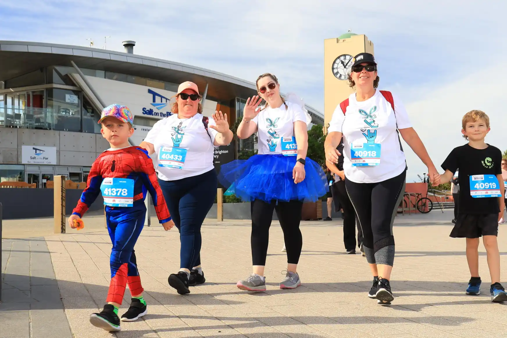 Three people are playing boccia in a gym. A young child stands to the left, preparing to throw. A young person with a prosthetic leg stands in the center, also preparing to throw. A person in a wheelchair holds a ball on the right. Balls are scattered on the court.