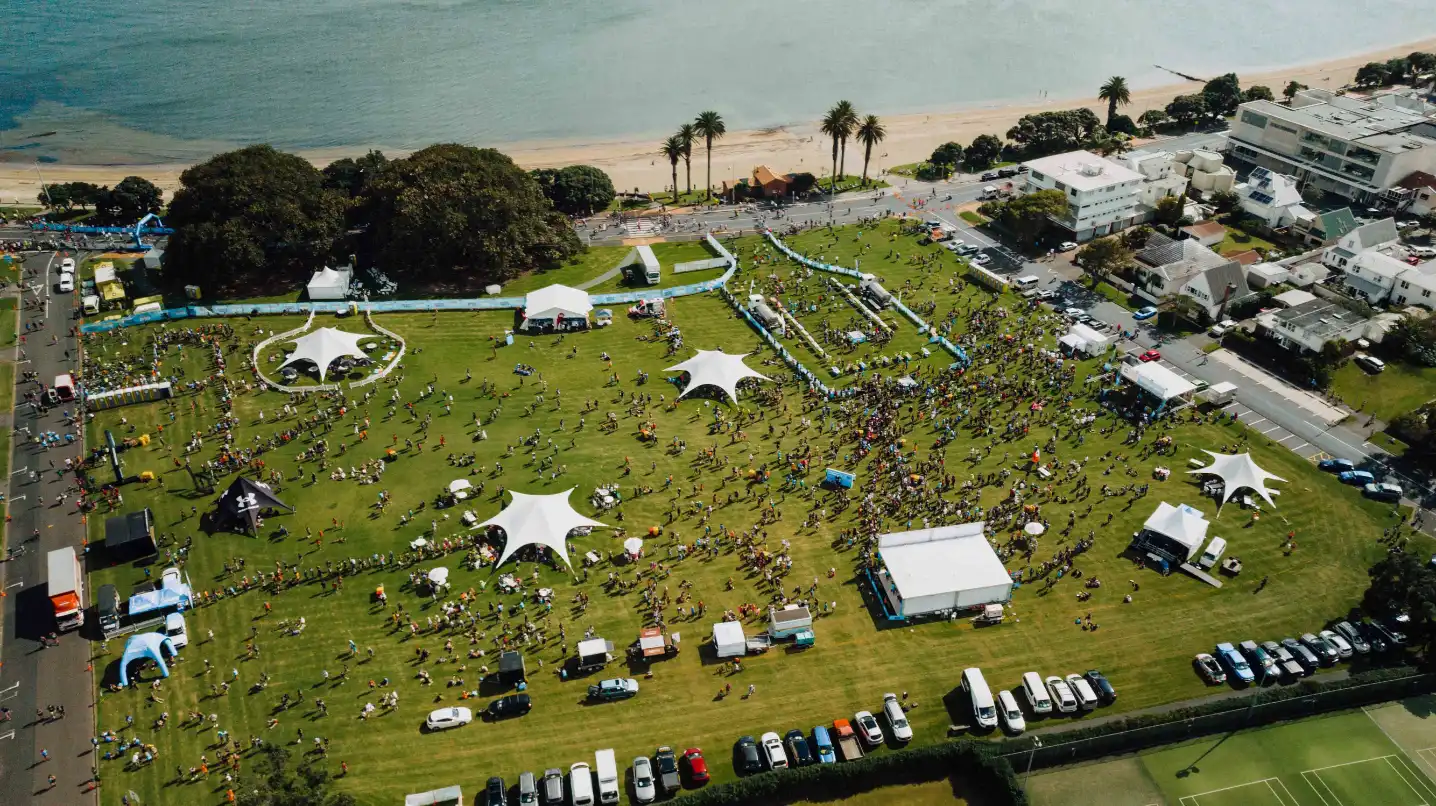 Aerial view of a seaside event with several tents, stalls, and people scattered across a grassy field. The event is near a beach with trees and parked cars visible. White tents and stages are set up, and people are mingling and enjoying the day.