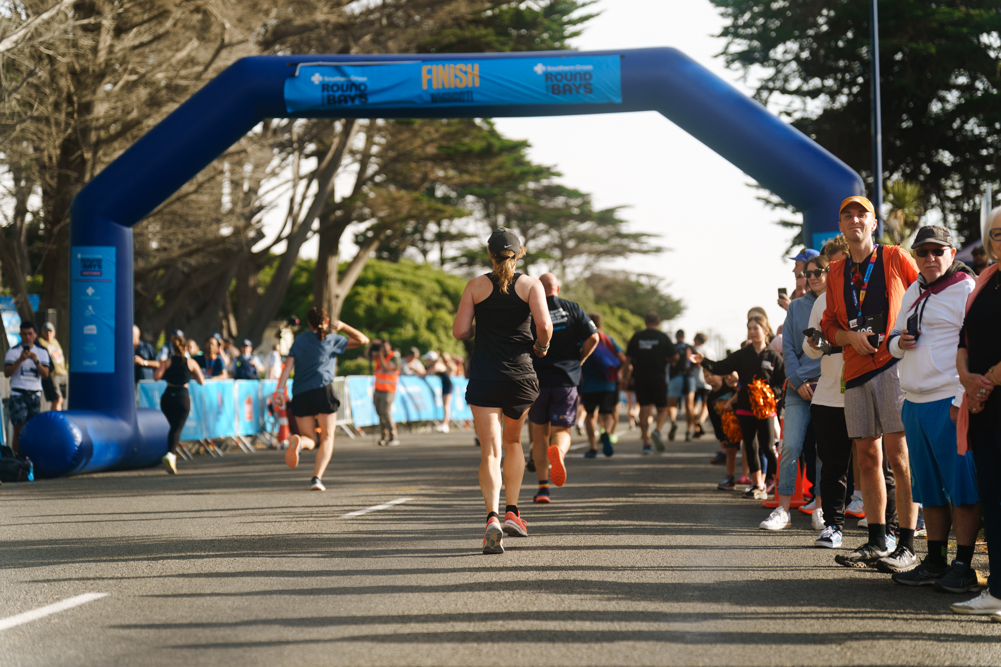 Three participants in yellow shirts celebrate with high-fives as they cross the finish line of a race. The finish line banner is blue and white. Spectators line both sides of the dirt track, cheering the runners. The sky is partly cloudy.