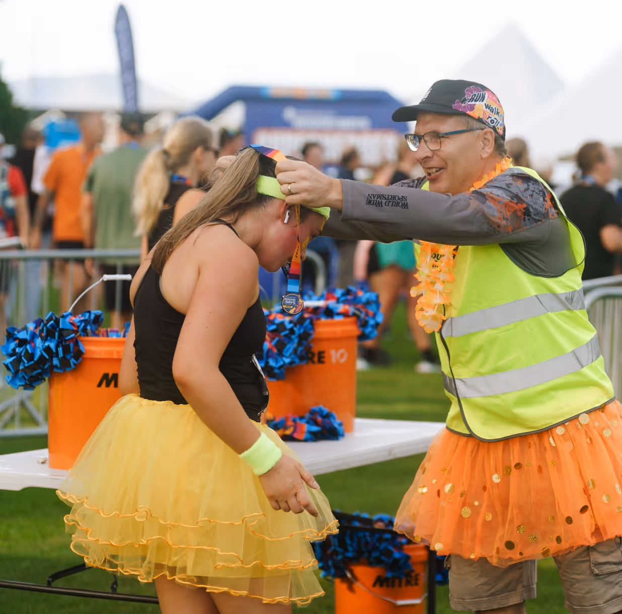 A runner crosses the finish line with arms outstretched in celebration. They are wearing a tank top, shorts, and running shoes. Spectators in the background cheer behind a blue barrier with the Under Armour logo. Trees and parked cars are visible behind the crowd.