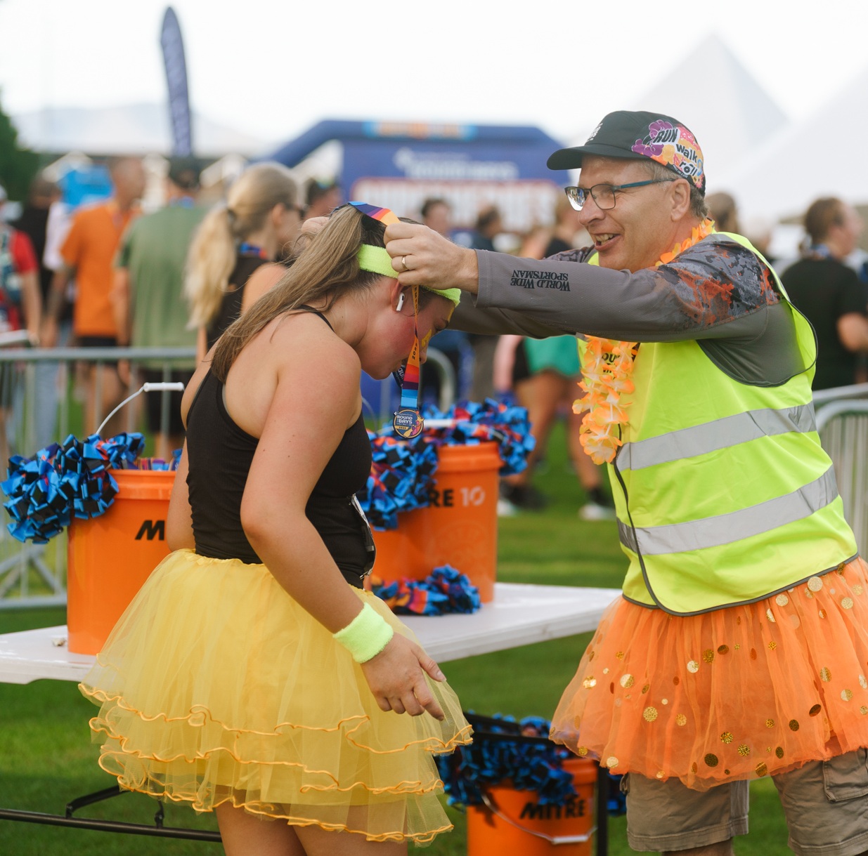 A runner crosses the finish line with arms outstretched in celebration. They are wearing a tank top, shorts, and running shoes. Spectators in the background cheer behind a blue barrier with the Under Armour logo. Trees and parked cars are visible behind the crowd.