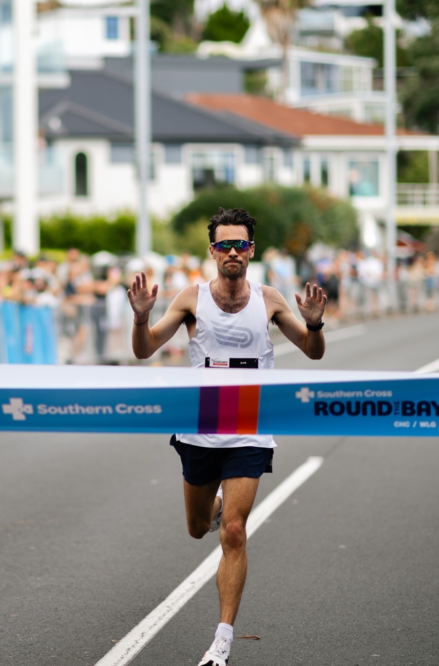 A female runner in a maroon outfit crosses the finish line at a race event sponsored by "Round the Bays." Spectators line both sides of the path, cheering her on as she passes under a blue inflatable arch. A person in a yellow vest stands to the right.