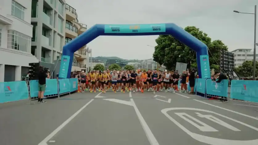 A large group of runners, including men and women, participates in a marathon along a seaside road. The sky is overcast, and there are hills and buildings visible in the distance across the water. The road has light posts, and some people watch from the sidelines.