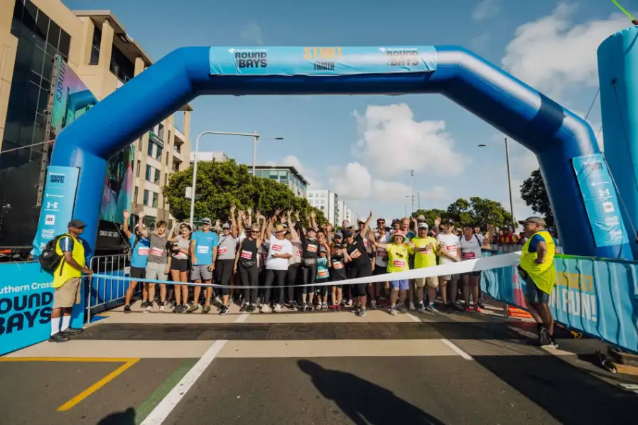 A large group of runners is gathered at the starting line of a race. The blue archway marking the "START" is prominently displayed. The scene is bustling with colorful attire and the road ahead is packed with participants. Trees and buildings are visible in the background.