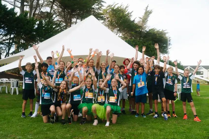 A group of children wearing yellow shirts and race bibs gather for a group photo outdoors. They appear to be participating in a charitable or sports event. The background features trees and buildings, and some participants are holding balloons.