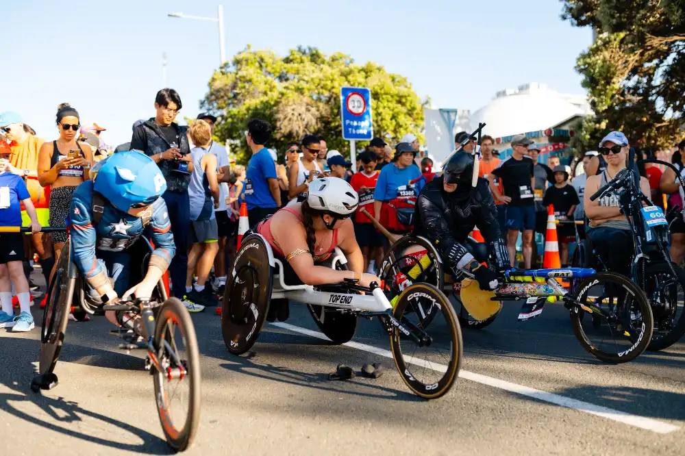 A group of people, including children and adults, are on a walk. Some of them are using wheelchairs. They are outdoors on a paved pathway with greenery and trees in the background. Everyone appears to be engaged and enjoying the walk.