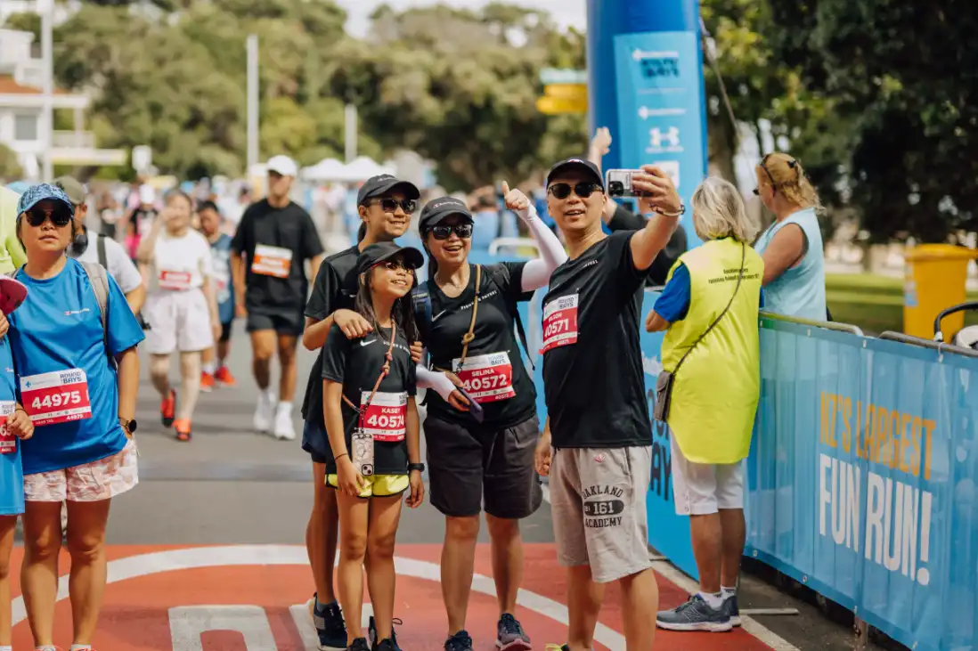 A group of runners poses for a photo at the finish line of a race event. Some participants are wearing race numbers and medals. A few other runners and spectators are seen in the background. The atmosphere is festive, with banners and flags around the area.